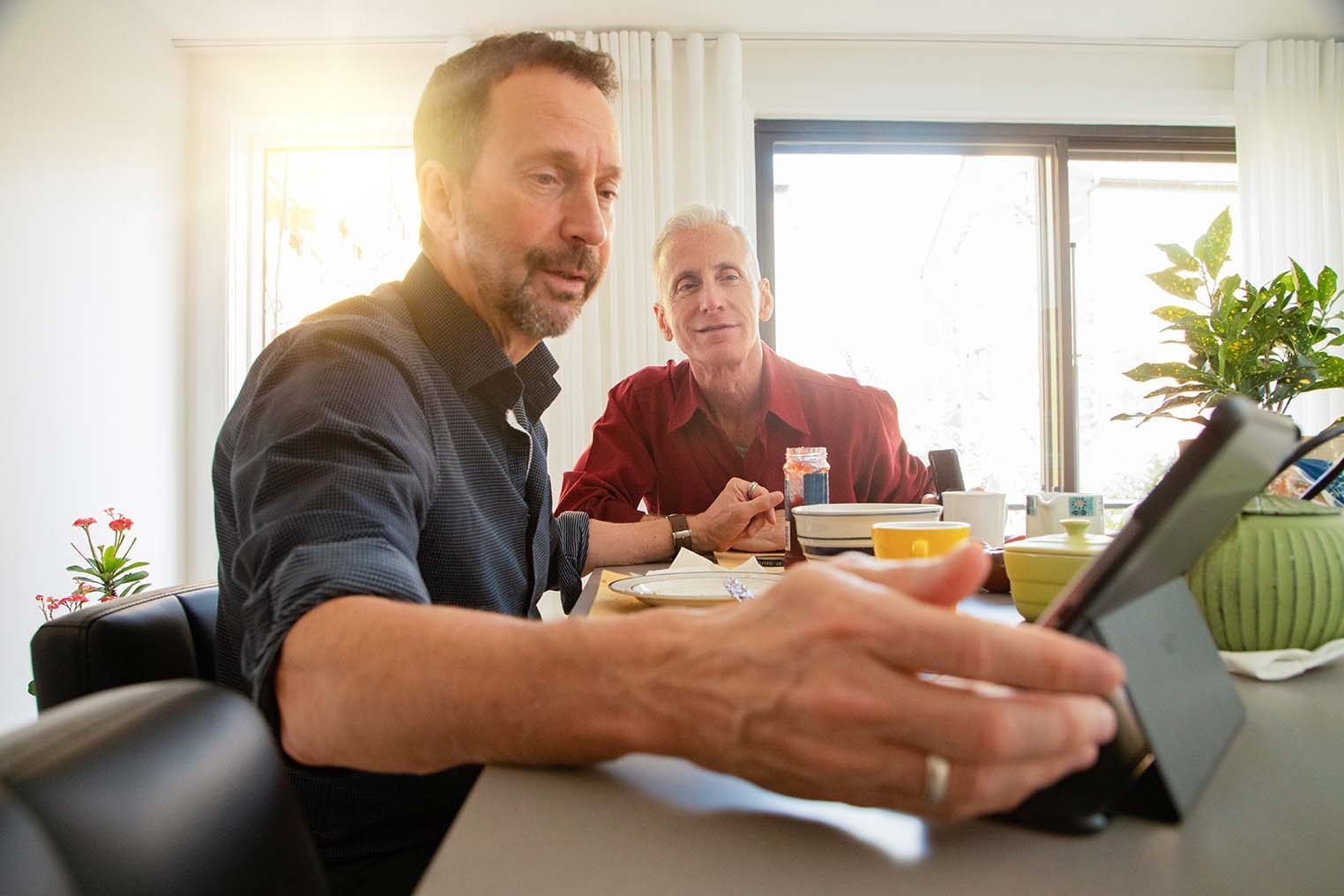 Senior gay couple having a lively conversation at breakfast about news on tablet